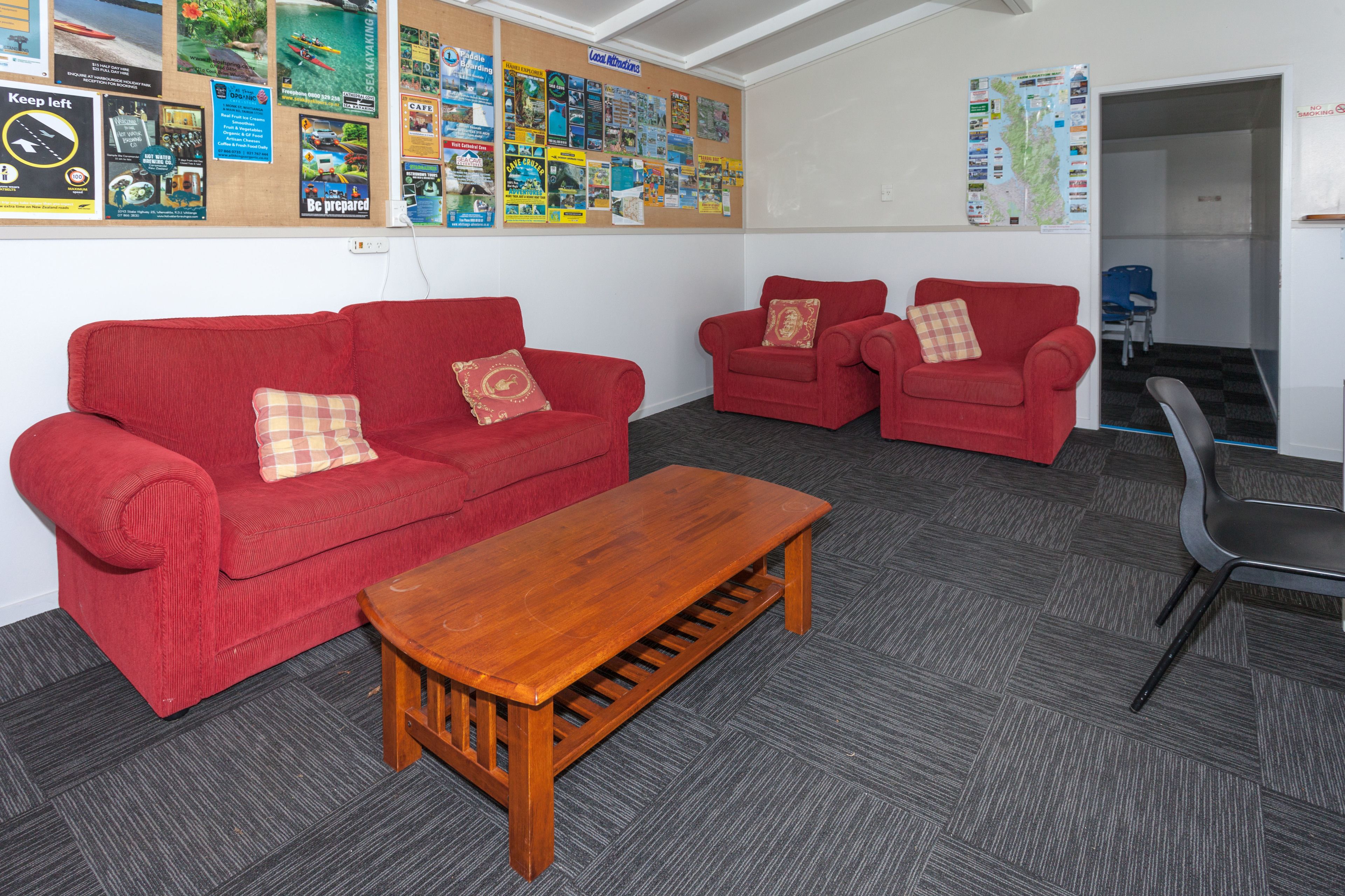 Quarto com dois sofás vermelhos, uma mesa de centro de madeira e um mural com diversos avisos.