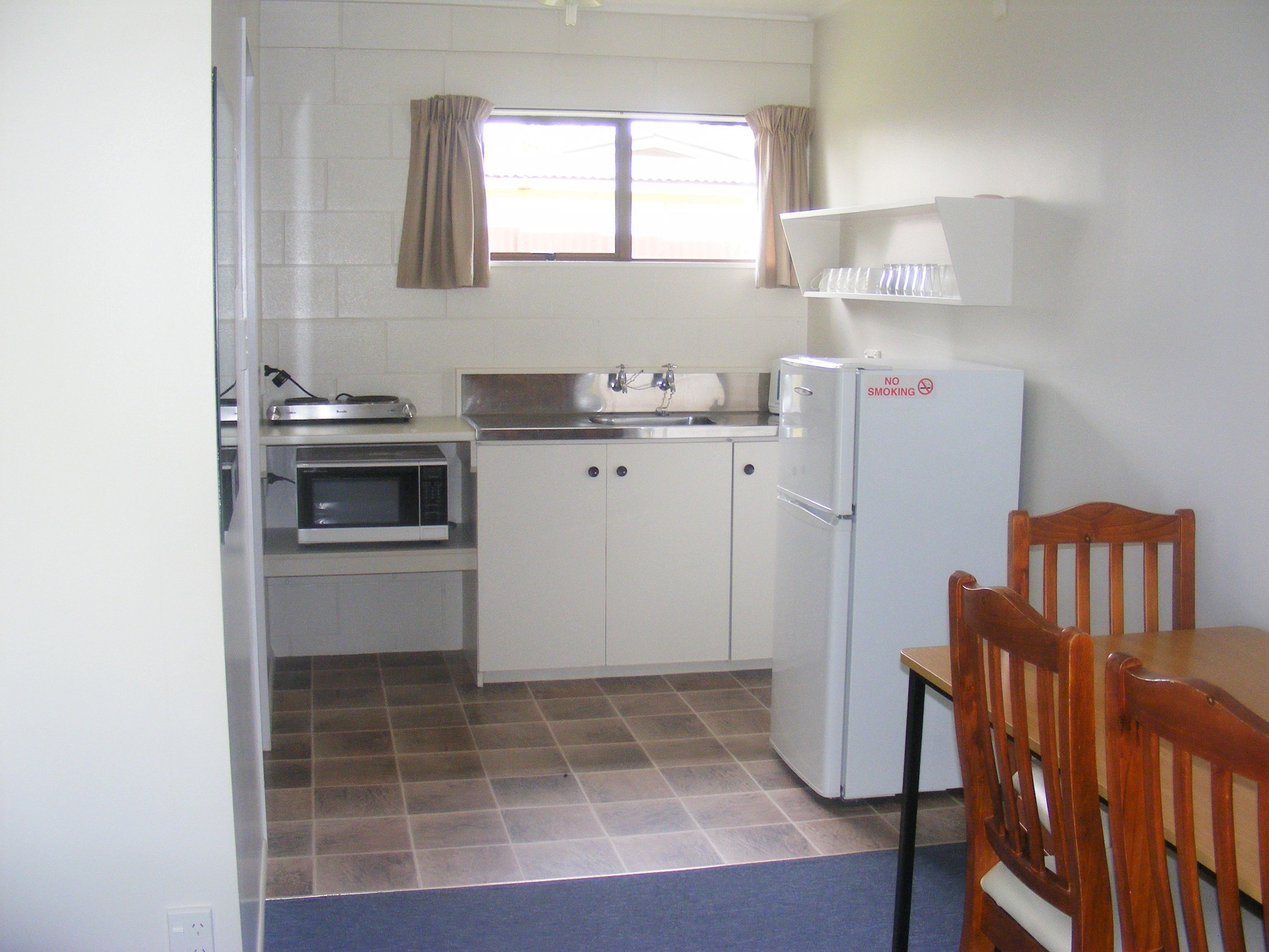 A compact kitchen with a microwave, oven, and refrigerator, a window with curtains, and a dining area with a wooden chair.