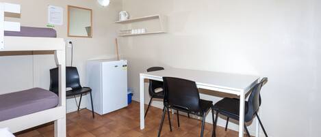 A dormitory room with bunk beds, a desk, two chairs, a refrigerator, and a shelf with items.