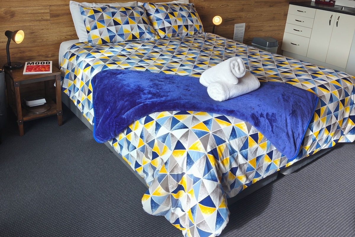 A hotel room with a wooden wall, a bed with a geometric patterned bedspread, a blue blanket, a white towel, a nightstand with a lamp, and a small shelf with a book.