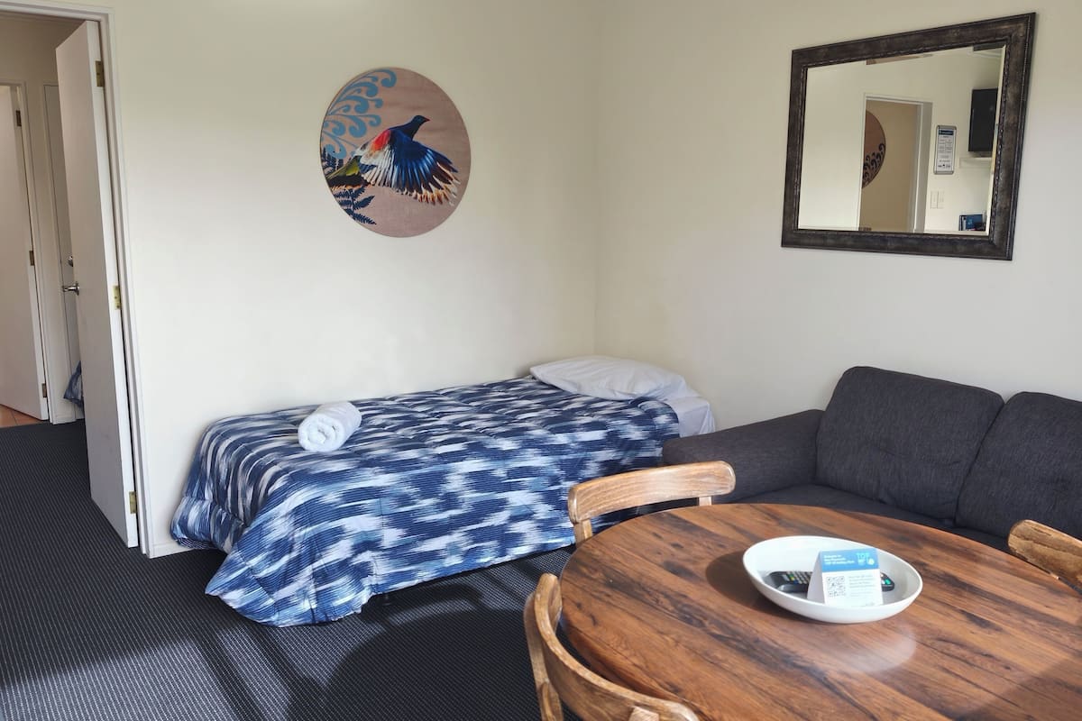 A room with a bed, a sofa, a wooden table, a mirror, and a wall art piece.
