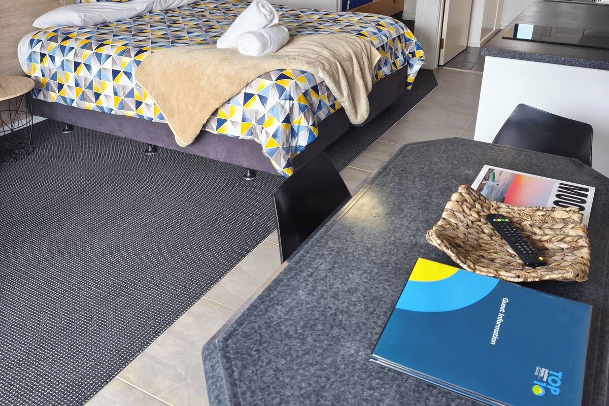 A hotel room with a bed, a desk, and a chair. There is a remote control on the desk and a brochure titled "Guest Information".