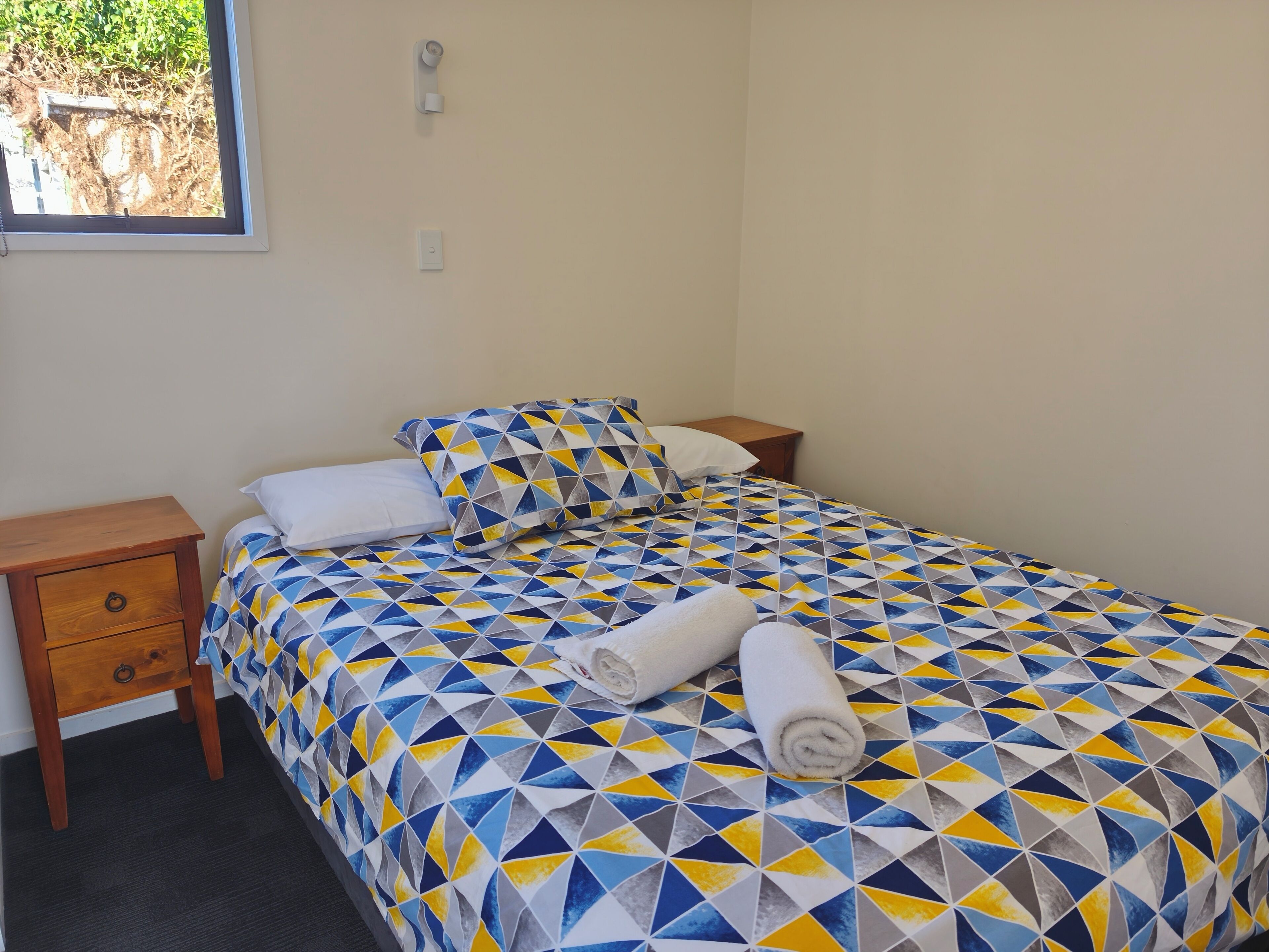 Una cama bien hecha con una colcha de estampado geométrico, una mesita de noche de madera con un cajón y una pequeña mesa al lado de la cama.