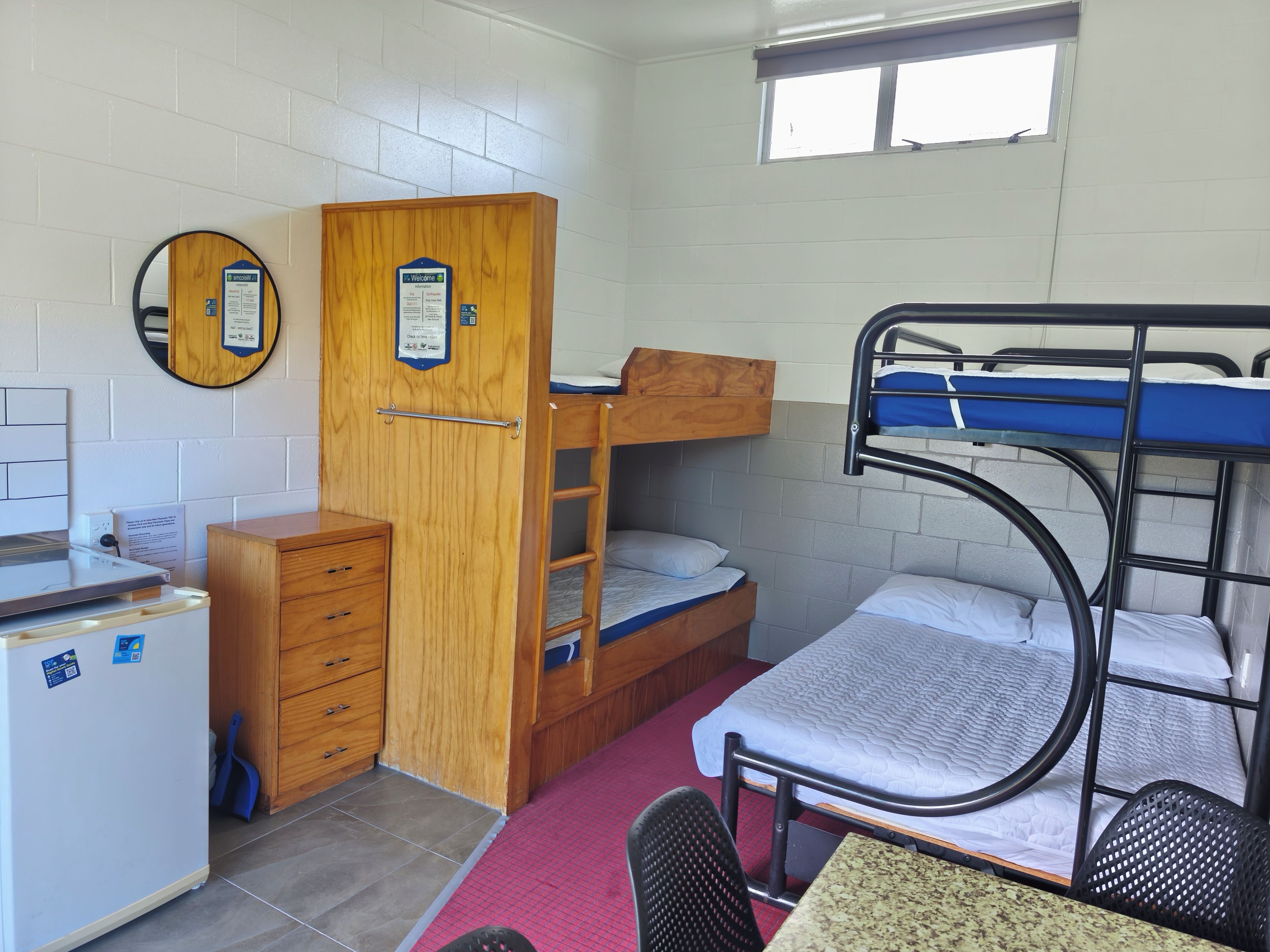 A dormitory room with bunk beds, a wooden wardrobe, a desk with a chair, and a small window.