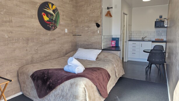 A bedroom with a bed, a bedside table, a wall-mounted artwork, and a view into a kitchen area with a microwave and a sink. - New Plymouth Top 10 Holiday Park (New Plymouth)