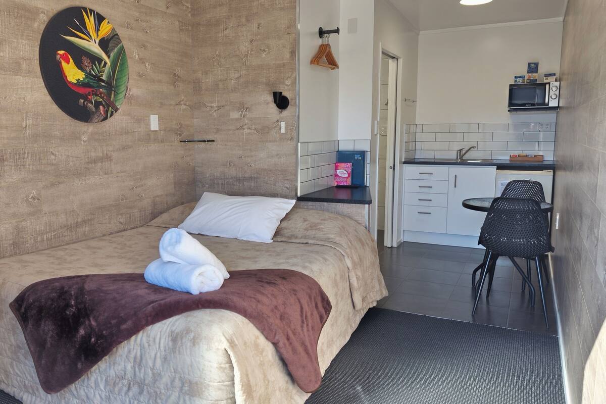 A bedroom with a bed, a bedside table, a wall-mounted artwork, and a view into a kitchen area with a microwave and a sink.
