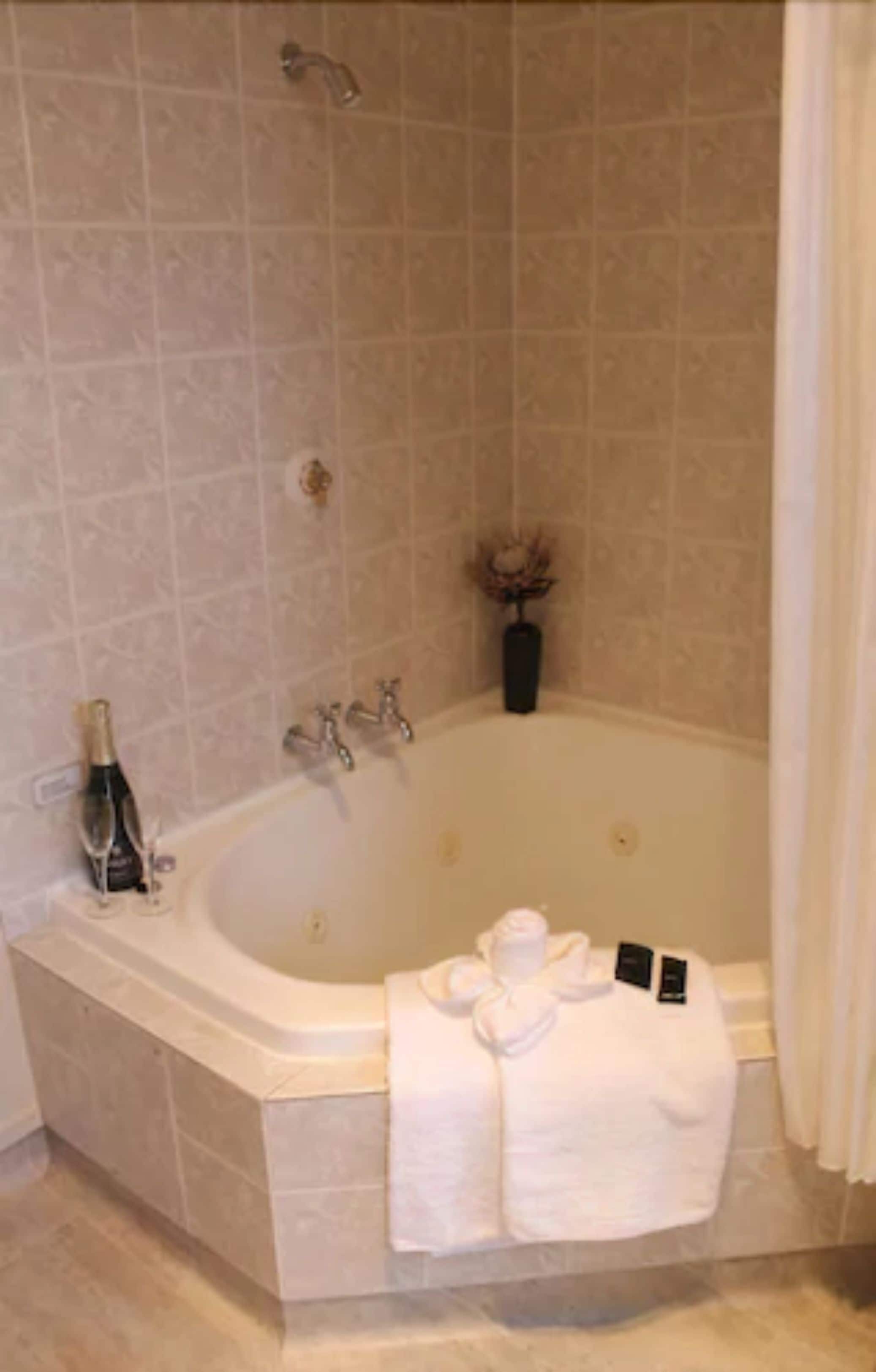 A large bathtub with a showerhead, a bottle of champagne, and a vase with dried flowers.