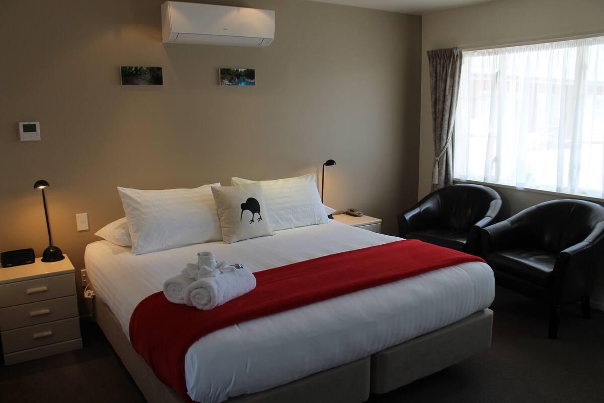A hotel room with a bed, a red blanket, a pillow with an animal design, a chair, and a window with curtains.