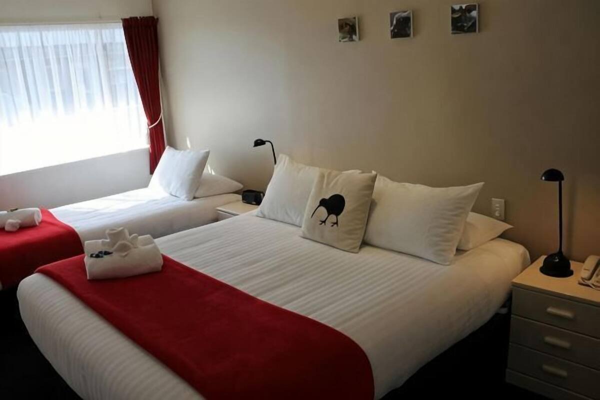 A hotel room with two beds, a red throw blanket, a white pillow with a black design, a bedside lamp, and a small table with a phone.