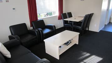 Una habitación con sofá, dos sillones, una mesa de centro, una mesa de comedor con sillas, un reloj en la pared y un ventanal con cortinas.