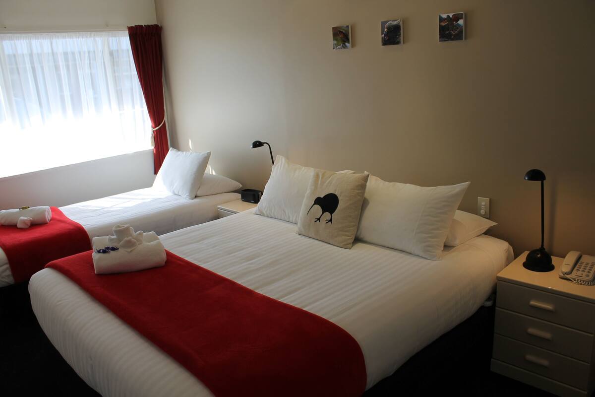 A hotel room with two beds, each with white bedding and red accents. There are wall-mounted picture frames, a bedside table with a lamp, and a telephone.