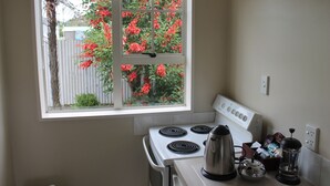 A kitchen with a stove, a coffee maker, and a window with red flowers outside. - Alpine Rose Greymouth Motel (Greymouth)