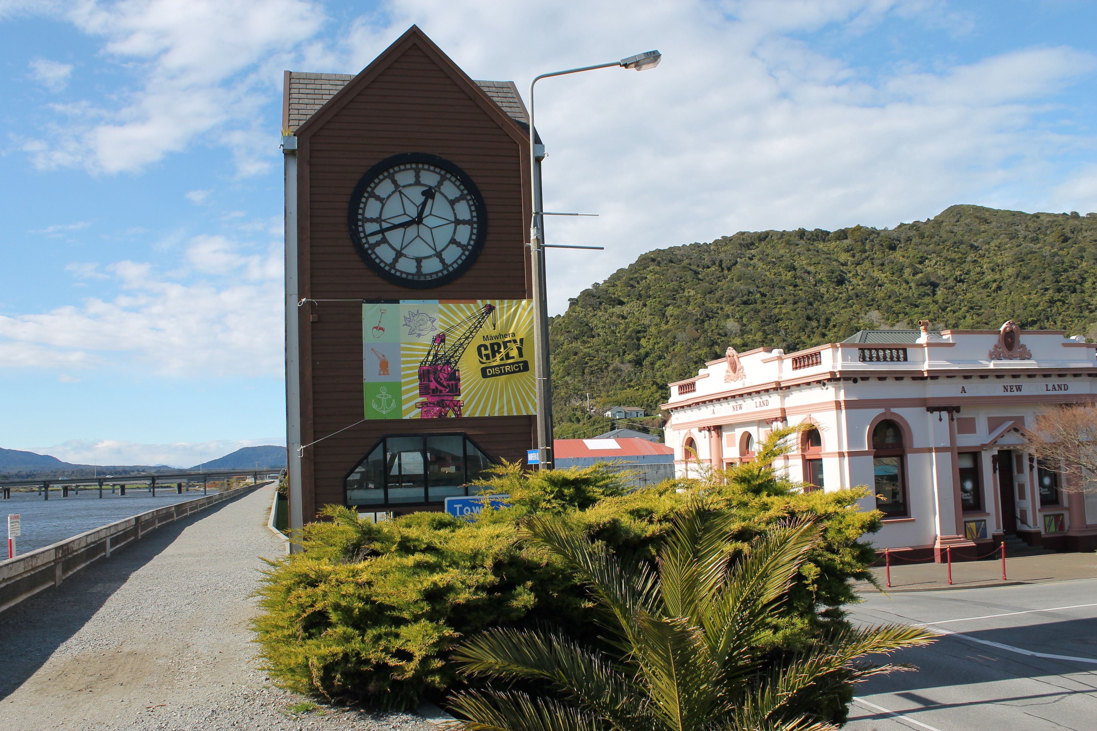 A building with a clock tower, a sign for Mawhera Grey District, and a view of a river and mountains in the background.