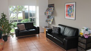 A modern living room with a black leather sofa, a coffee table, and a potted plant. There is a window with a view of the outside, and a wall-mounted information board. - Alpine Rose Greymouth Motel (Greymouth)