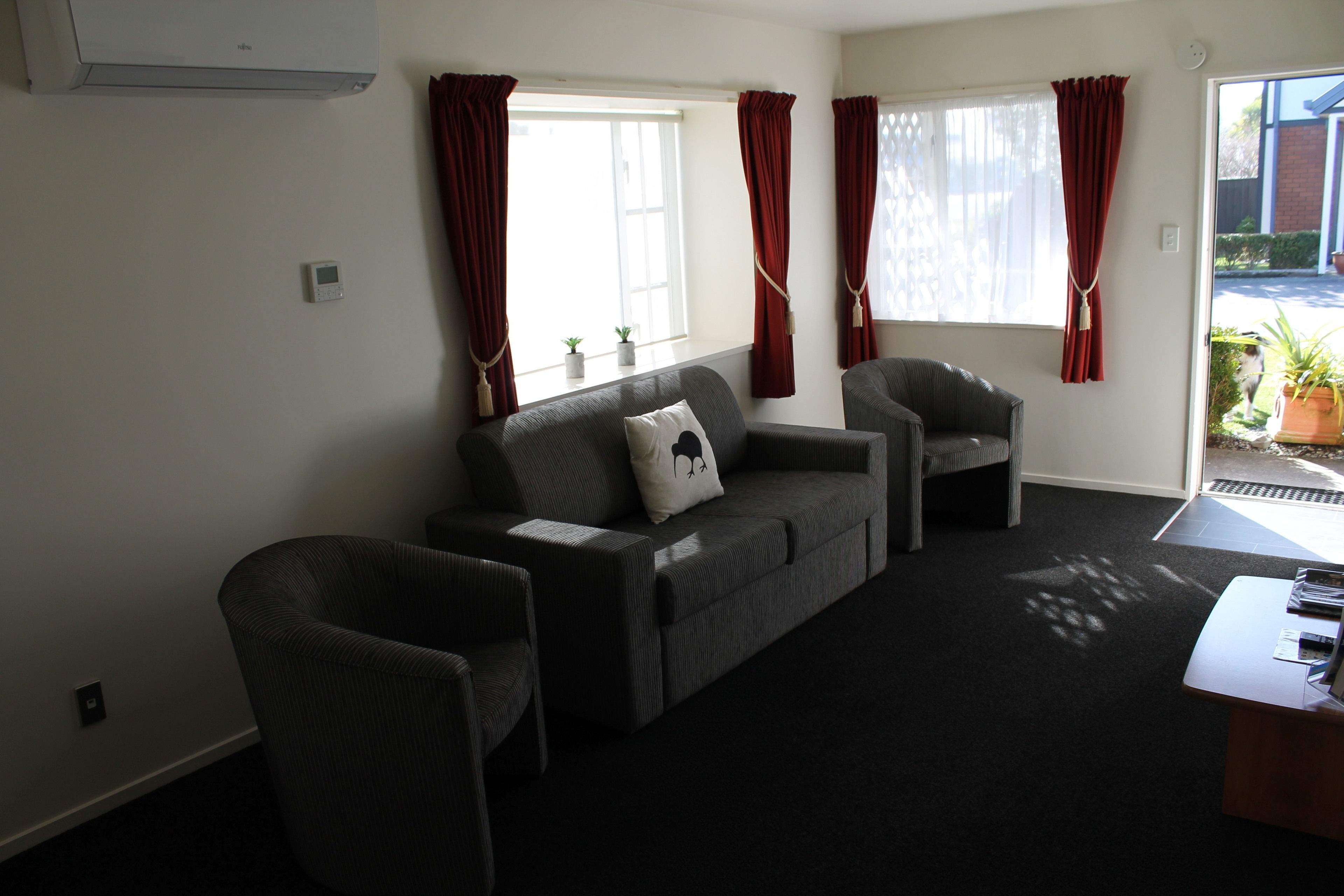 A living room with a sofa, armchair, and a small table. There are two windows with red curtains, a potted plant on the windowsill, and a door leading outside.