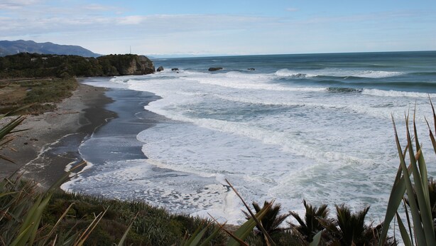 A coastal landscape with waves crashing onto the shore, a clear sky, and distant mountains. - Alpine Rose Greymouth Motel (Greymouth)