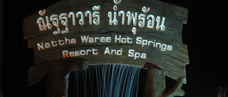 Une enseigne en bois, avec une inscription en thaï et un texte en anglais, indiquant l’emplacement du complexe hôtelier et du centre de bien-être Nattha Waree Hot Springs.