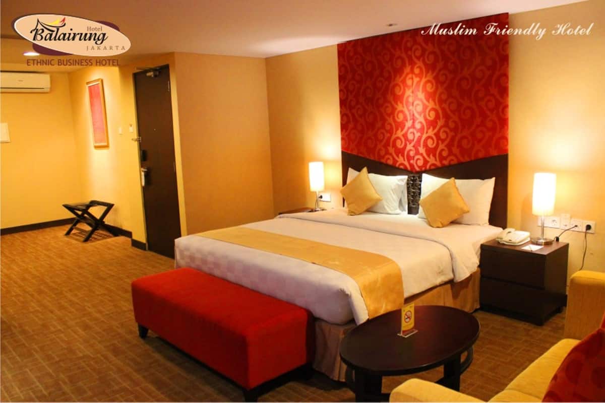 A hotel room with a large bed, a red bench, a small table, and a nightstand with a phone.