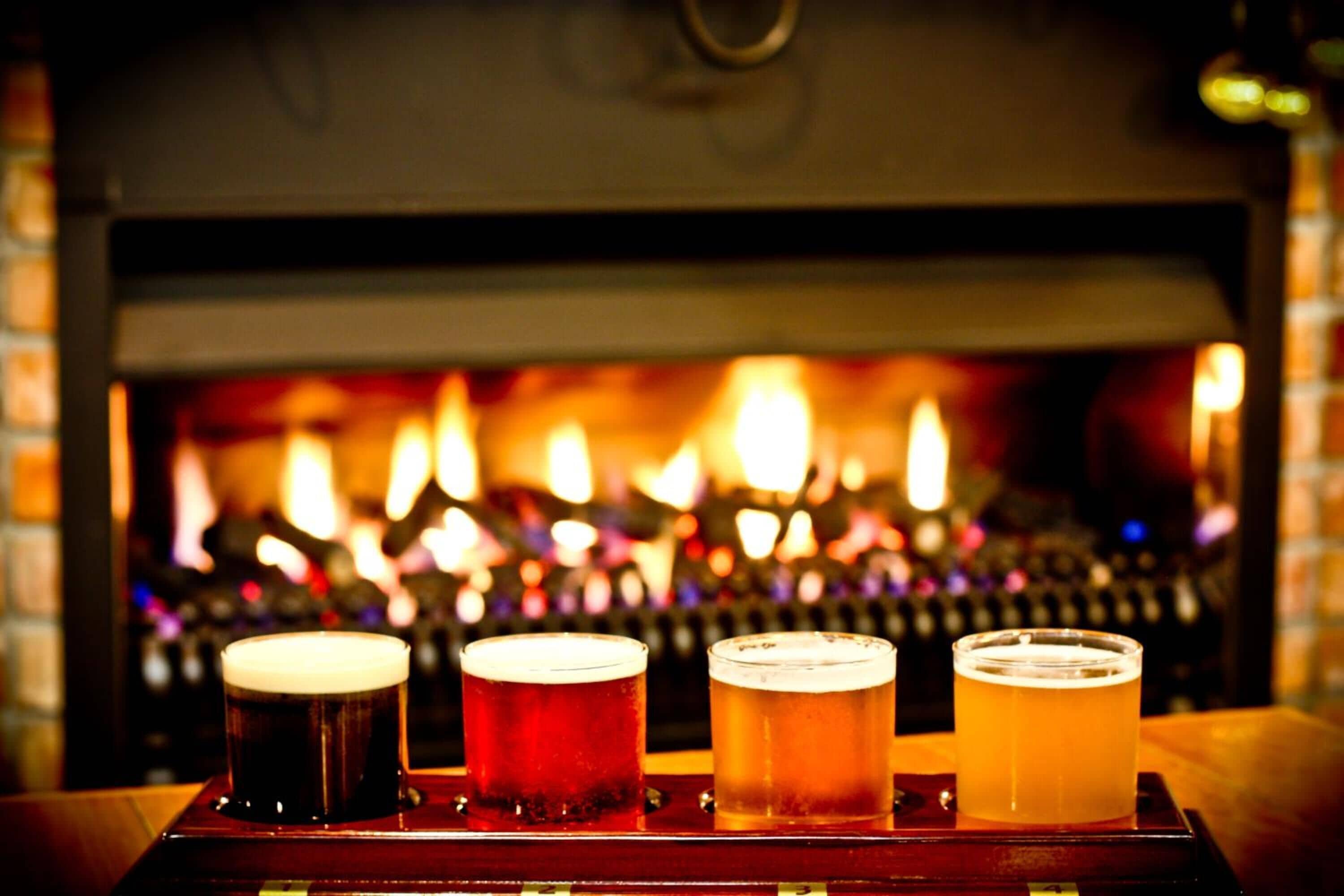 Cuatro vasos de cerveza sobre una bandeja frente a una chimenea con pared de ladrillo y rejilla metálica.