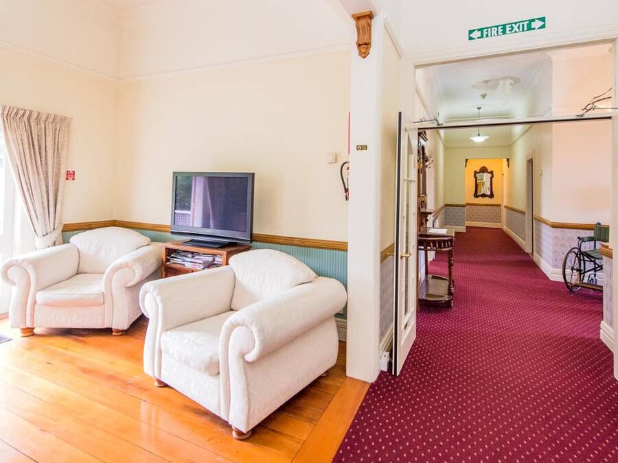 A hotel room with two white armchairs, a television, and a red carpeted hallway leading to a door marked "Fire Exit".