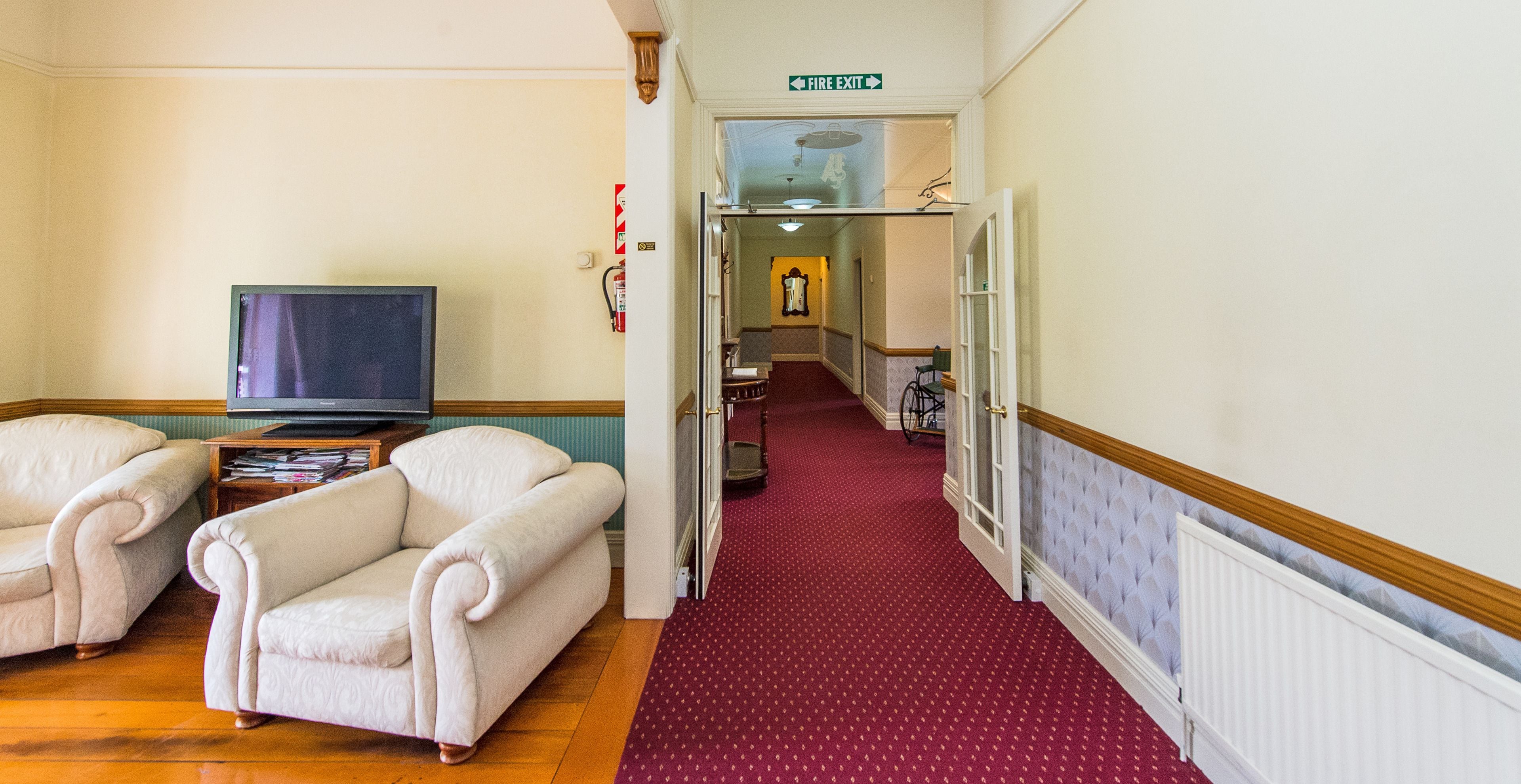 A hallway with a red carpet, a television on a stand, two armchairs, and a fire exit sign.