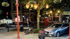 A restaurant with a signboard, a car parked nearby, and a tree with hanging lights.