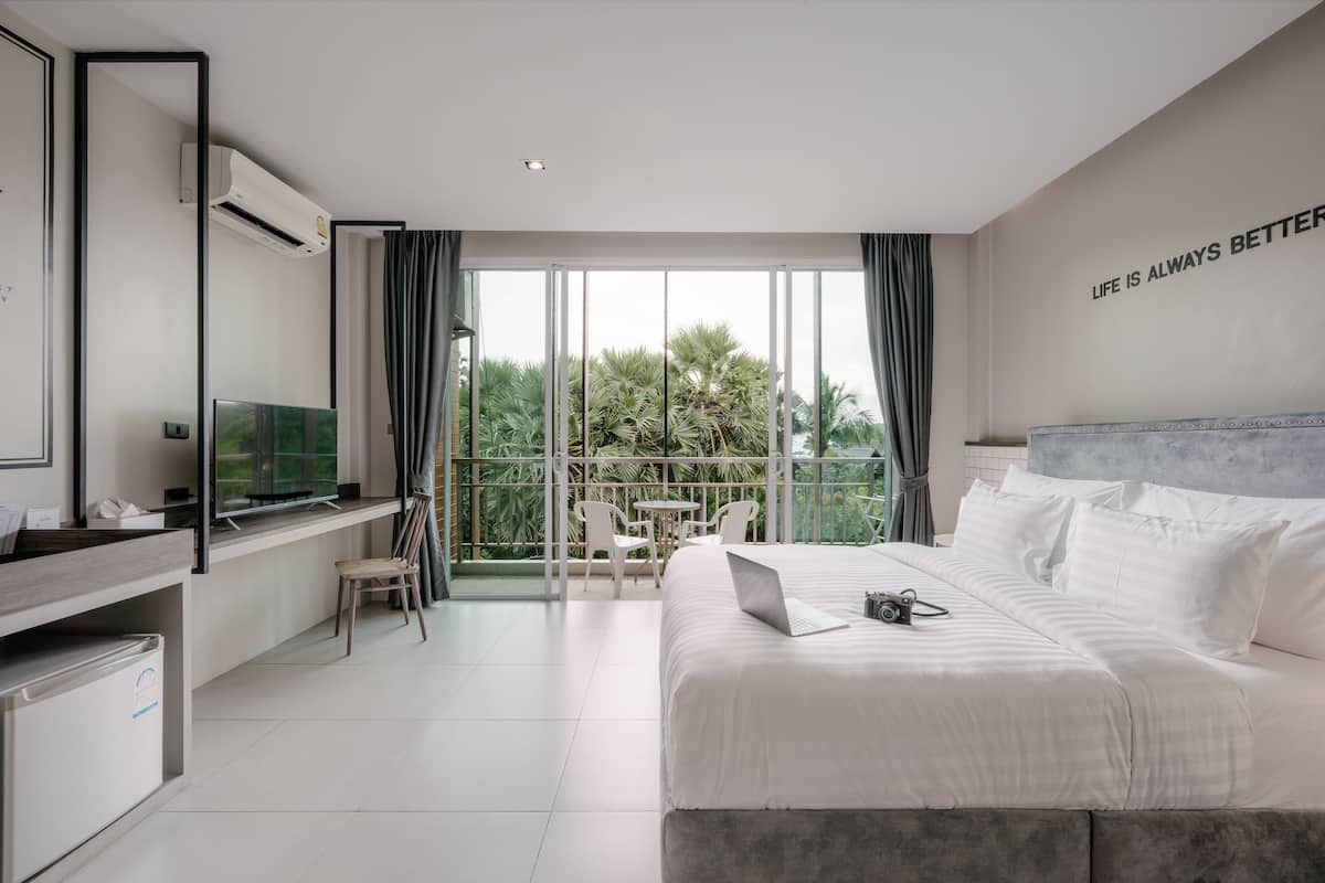 A modern hotel room with a large bed, a desk with a laptop, a TV, and a balcony with a view of greenery.