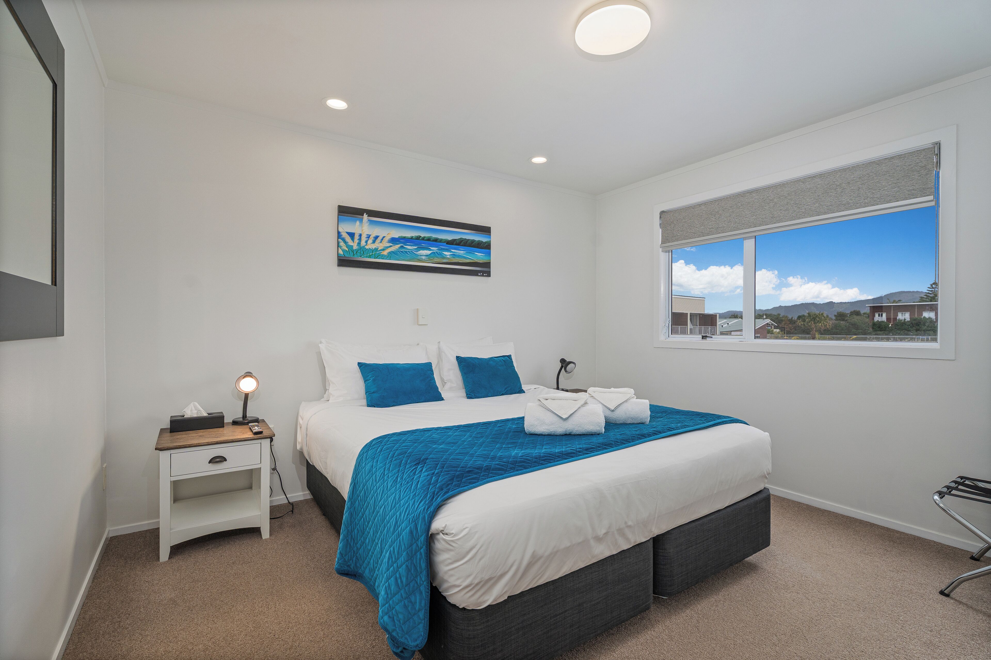 A modern bedroom with a large bed, a nightstand, a painting, and a window with a view.