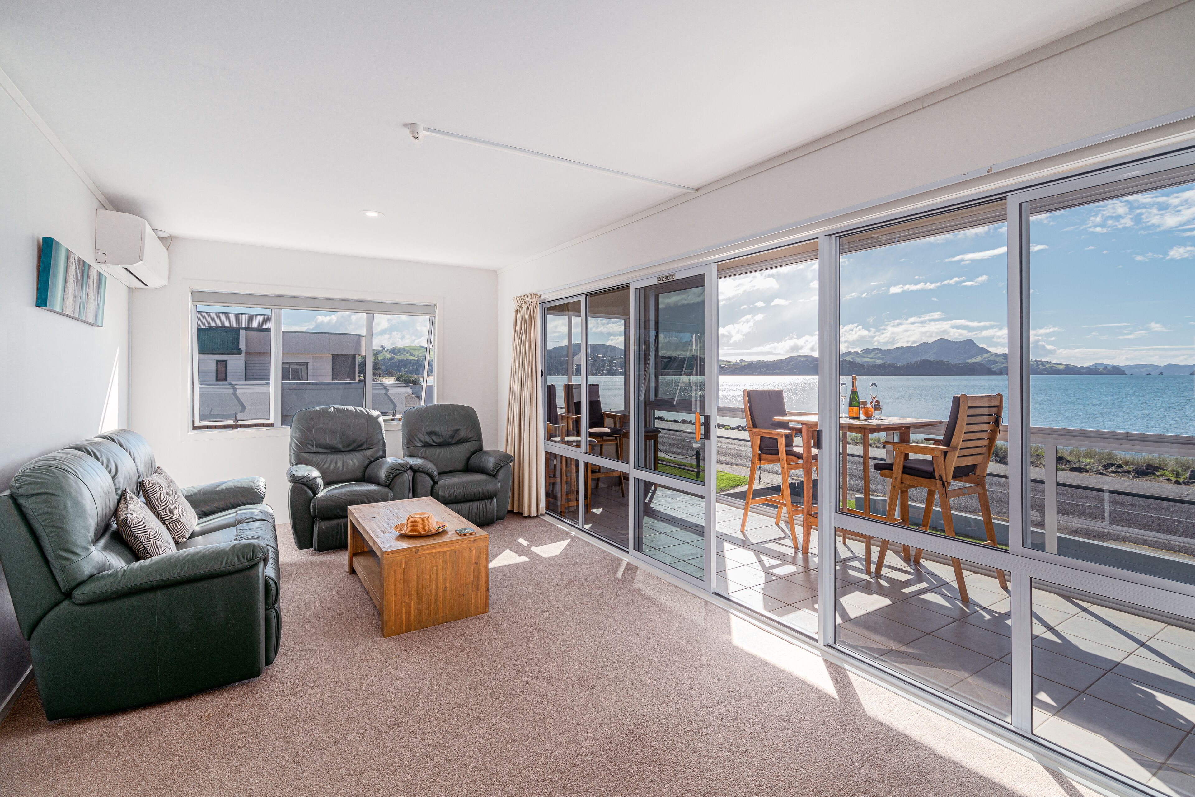 A modern living room with a green sofa, two armchairs, a wooden coffee table, and a large sliding glass door leading to an outdoor seating area with a table and chairs.