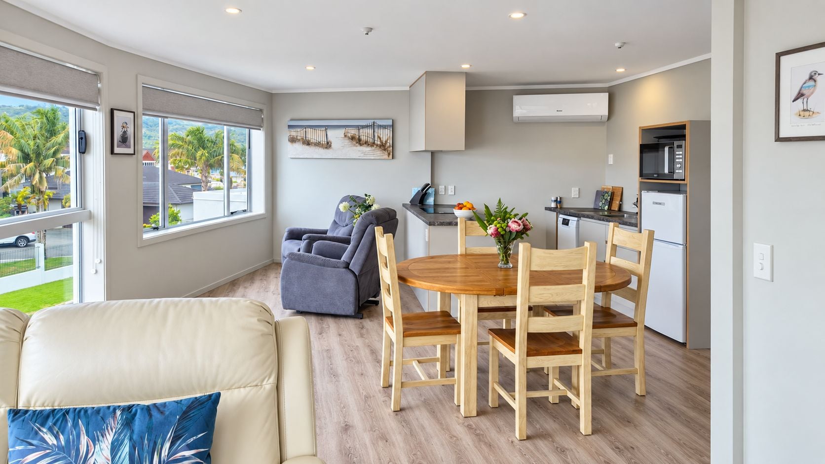 En moderne stue med sofa, spisebord med stole, en grå lænestol og et køkken med hårde hvidevarer.
