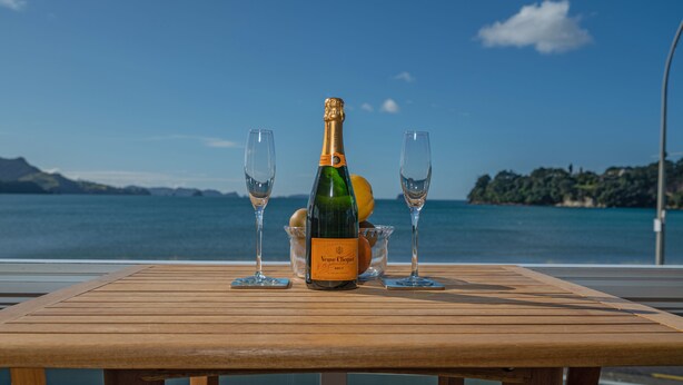 Una mesa de madera con una botella de Veuve Clicquot Brut champagne, dos copas y un cuenco de fruta frente a una vista panorámica del lago.
