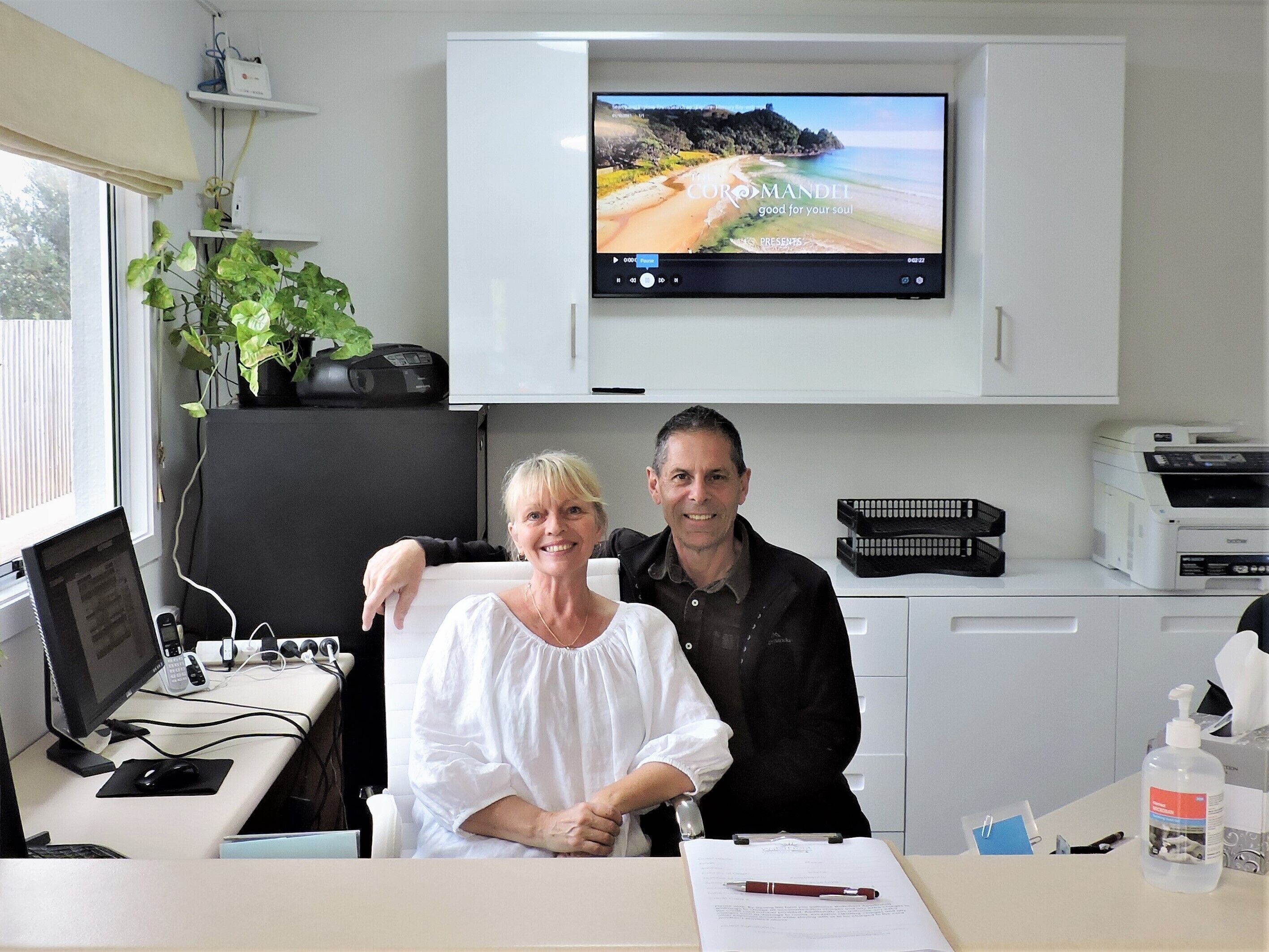 A room with a desk, computer, and a television displaying a beach scene.