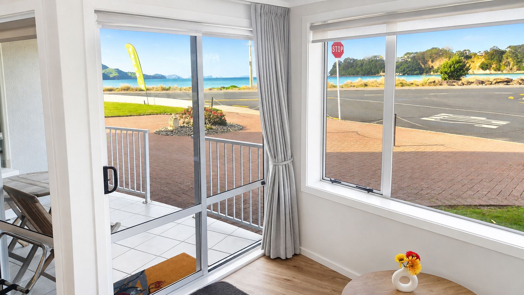 Et værelse med udsigt over en parkeringsplads og et stopskilt, en balkon med bord og stole, samt et lille bord med en vase blomster.