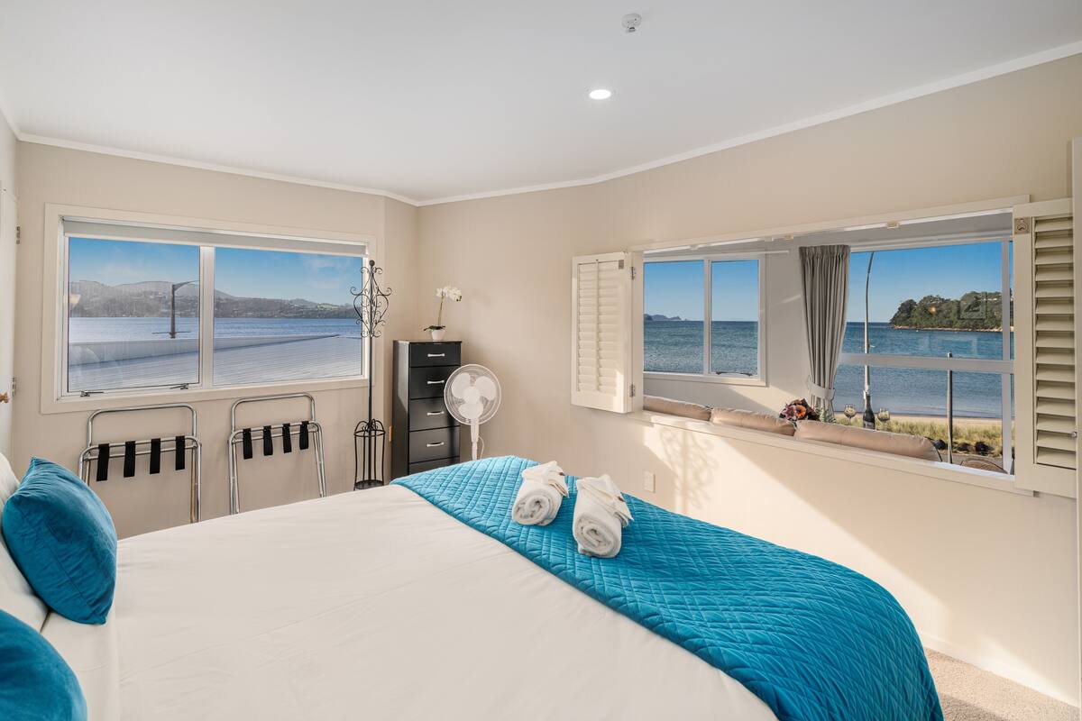A bedroom with a large bed, two windows with ocean views, a fan, and a dresser.