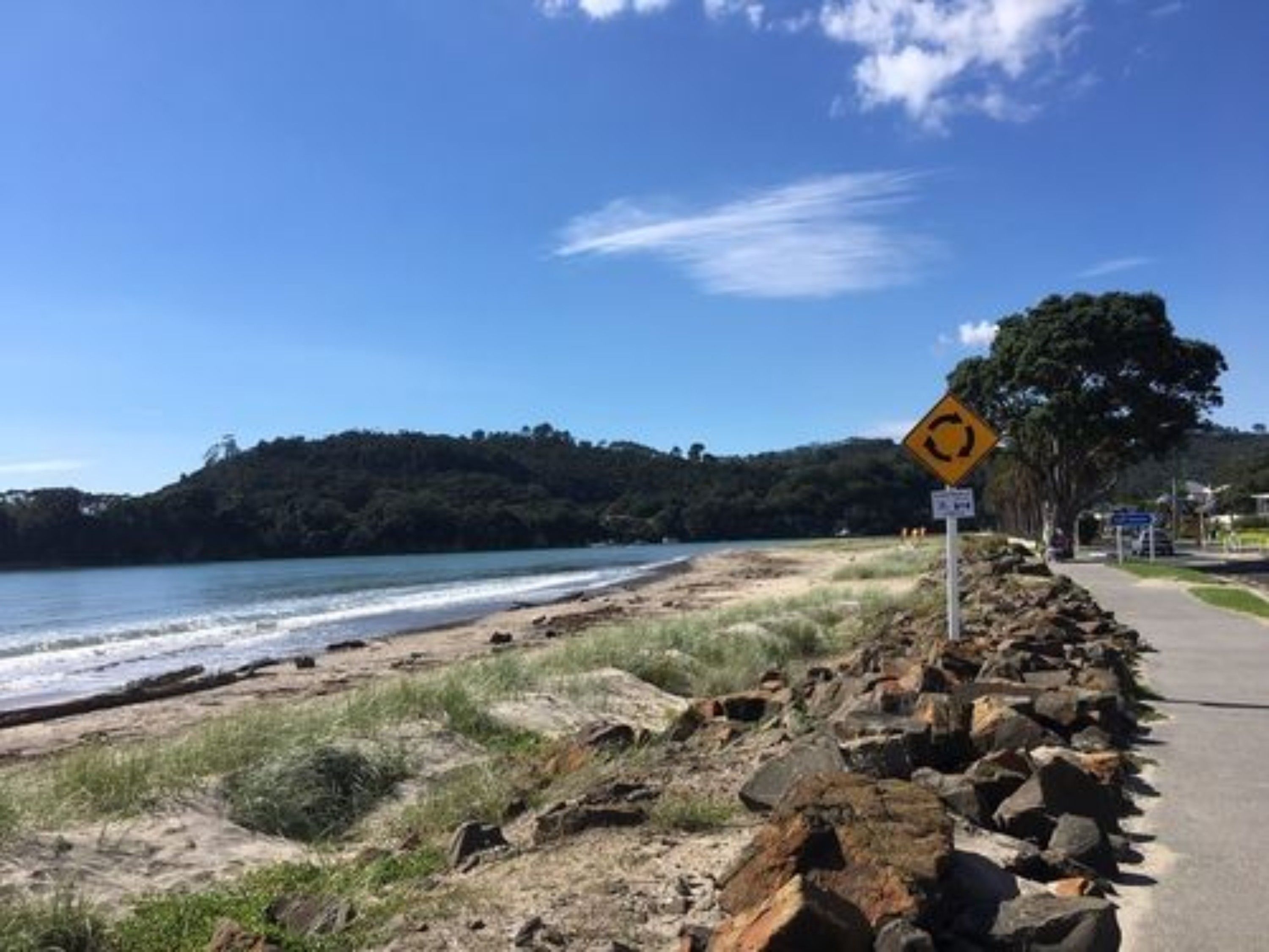 Une route côtière, agrémentée d’un panneau d’avertissement et d’une barrière rocheuse, offrant une vue dégagée sur la mer et les collines en arrière-plan.