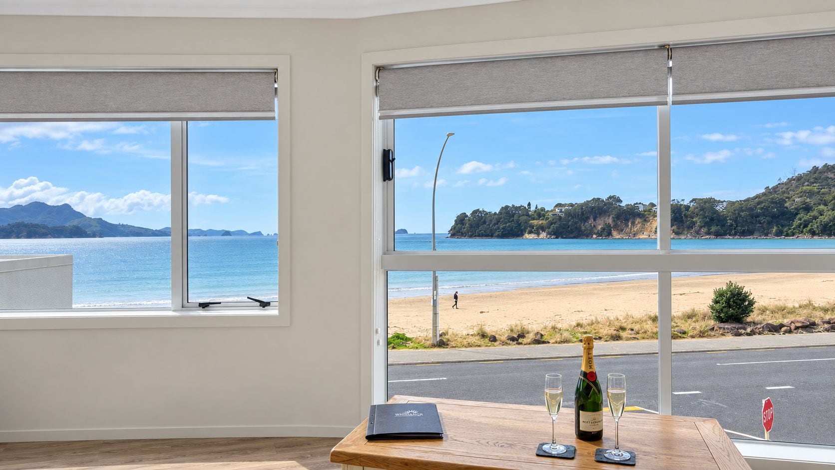 Een kamer met grote ramen met uitzicht op een strand, een houten tafel met een fles en glazen, en een laptop.