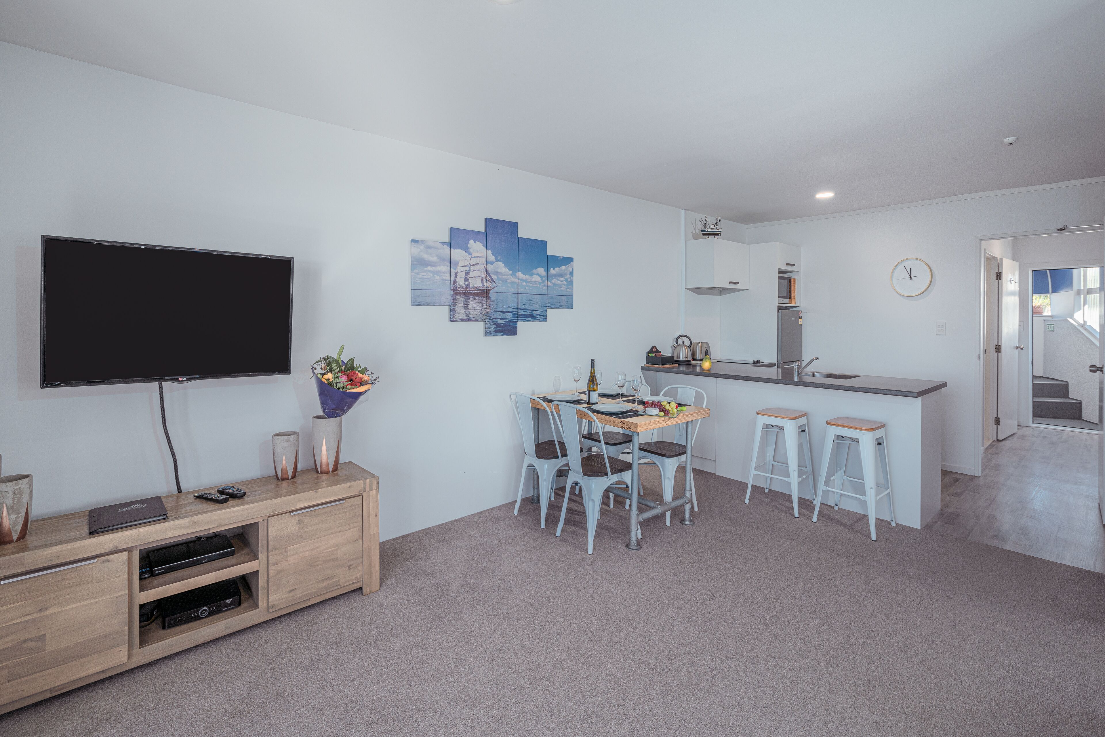 Una sala de estar moderna con televisor de pantalla plana, mueble de madera para entretenimiento, mesa de comedor con cuatro sillas y una zona de cocina con fregadero y encimera.
