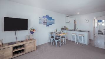 A modern living room with a flat-screen TV, wooden entertainment unit, dining table with four chairs, and a kitchen area with a sink and countertop.