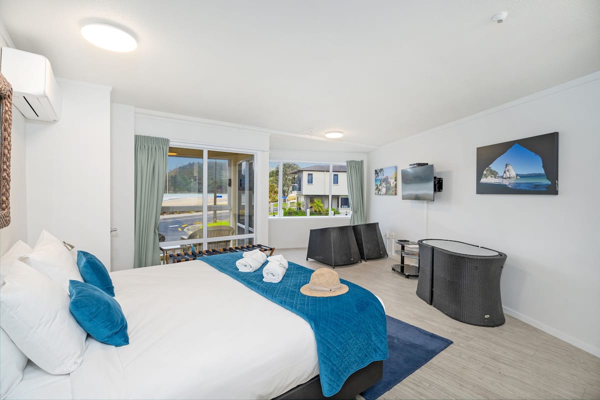 A hotel room with a large bed, a view of the outside, a small table with chairs, and a trash bin.