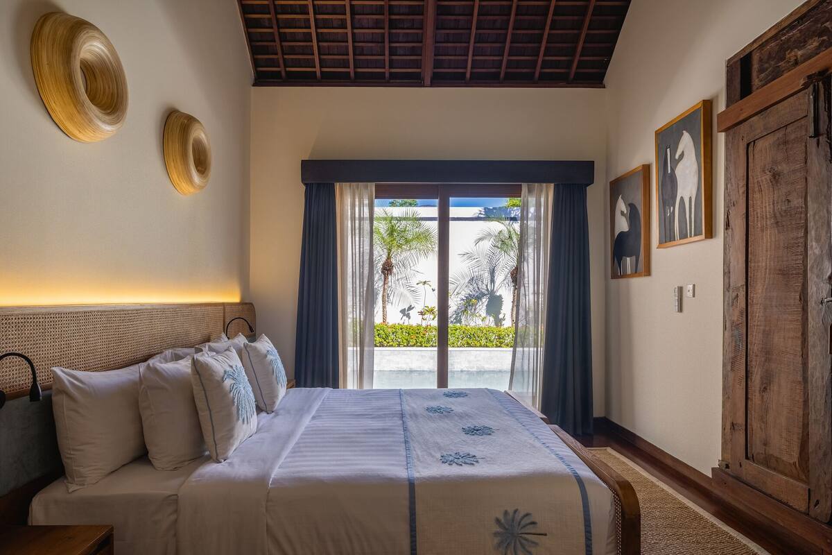A bedroom with a bed, a wooden headboard, a large window with curtains, and two wall-mounted decorative items.