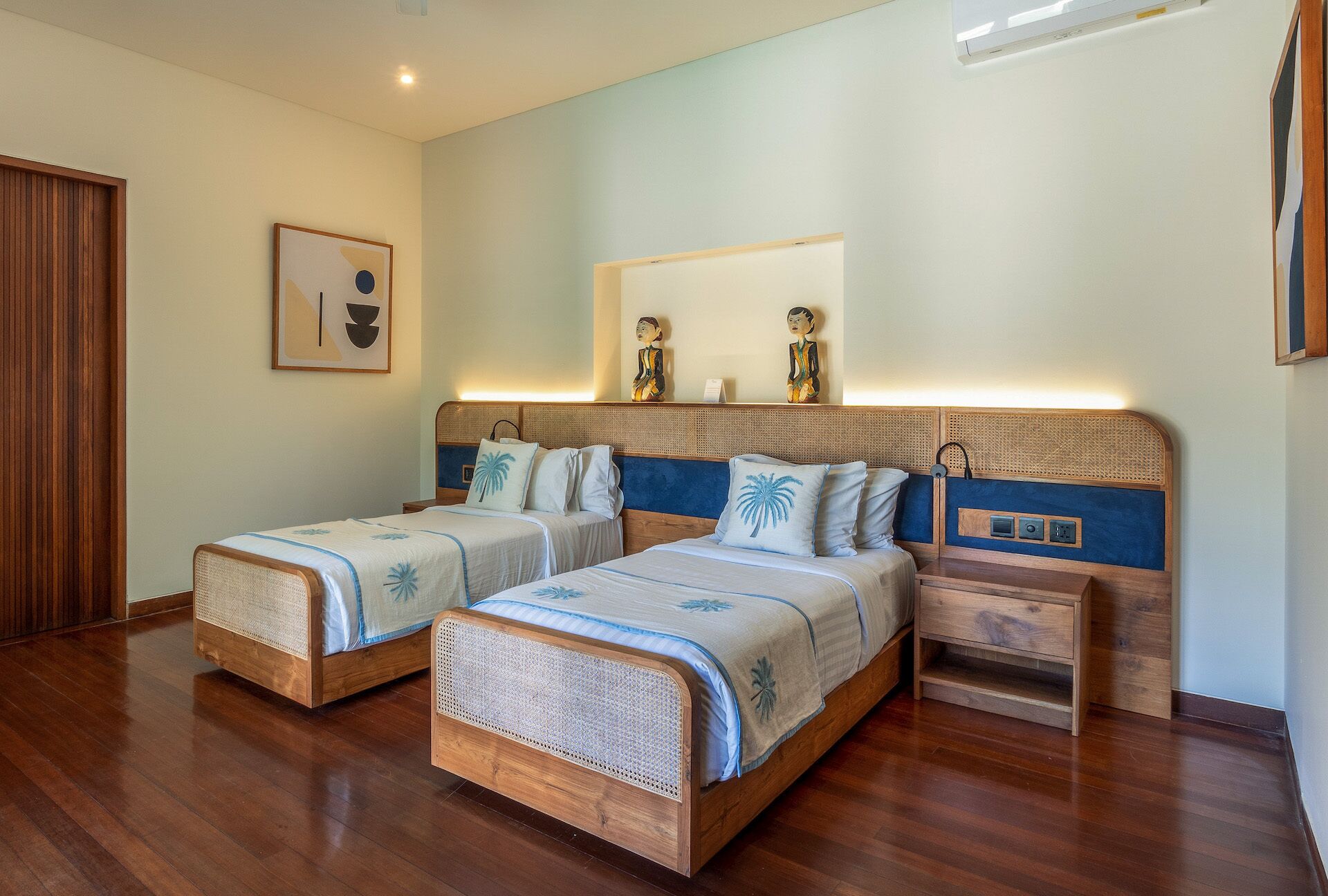 Quarto duplo com duas camas de madeira, uma mesinha de cabeceira, um espelho fixado na parede e um quadro emoldurado.
