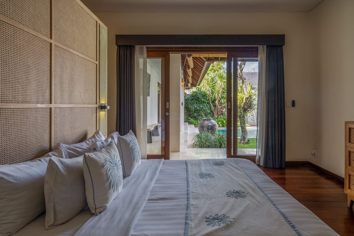 A bedroom with a large bed, a woven headboard, and a view of a garden and pool through a sliding glass door.