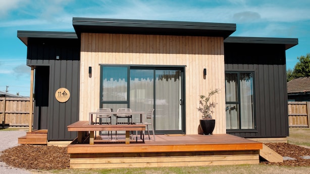 Een modern huis met een houten terras, een eethoek met stoelen en een grote schuifpui.
