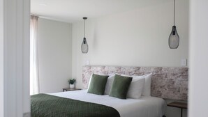 Een net opgemaakt bed met een groene deken, twee groene kussens en een wit hoofdbord. Daarboven hangen twee hanglampen.