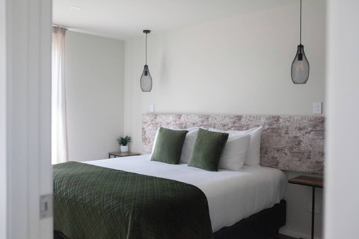Een net opgemaakt bed met een groene deken, twee groene kussens en een wit hoofdbord. Daarboven hangen twee hanglampen.