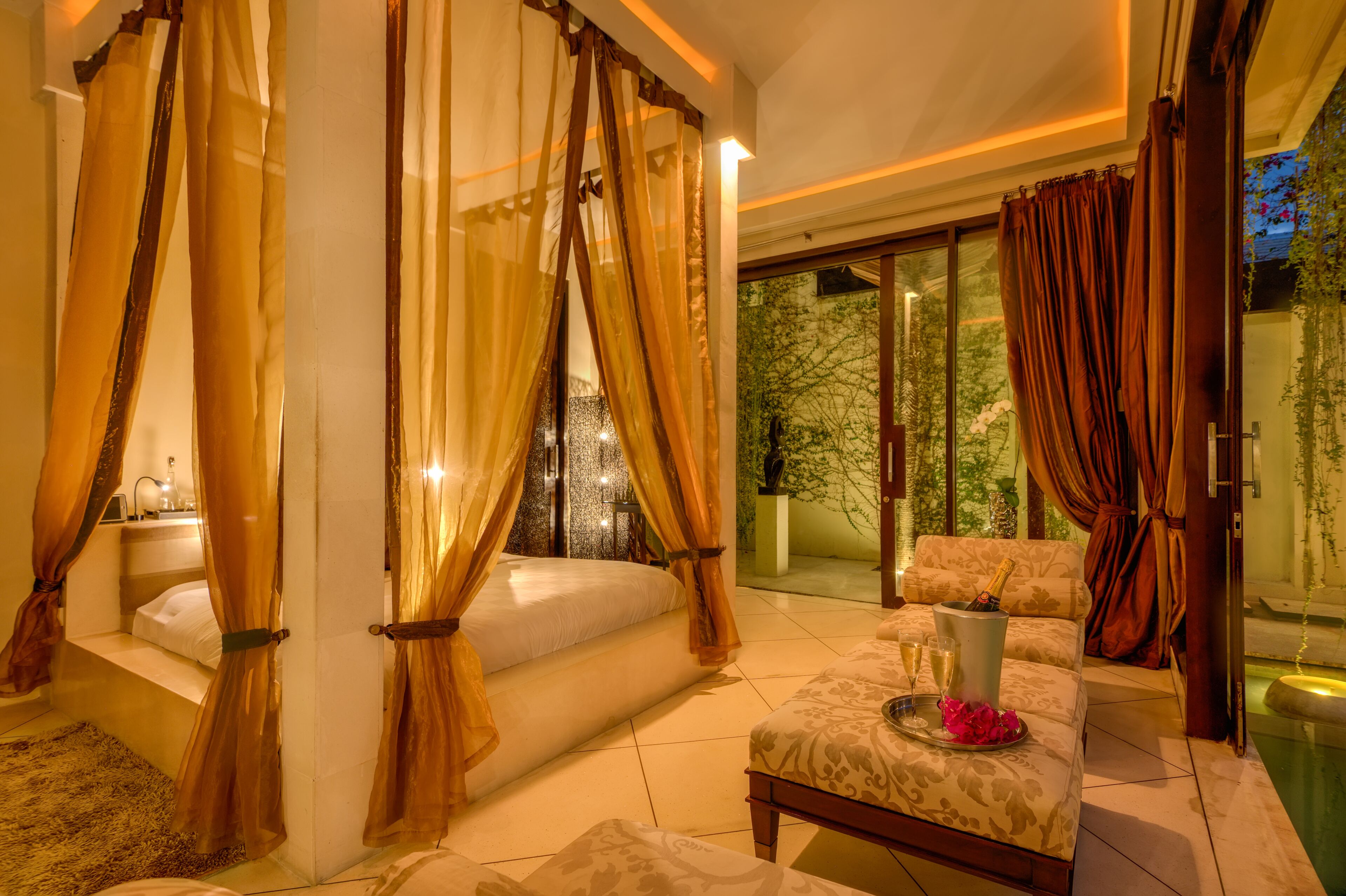 A luxurious bedroom with a canopy bed, a sitting area with a champagne bottle, and a view of greenery outside.