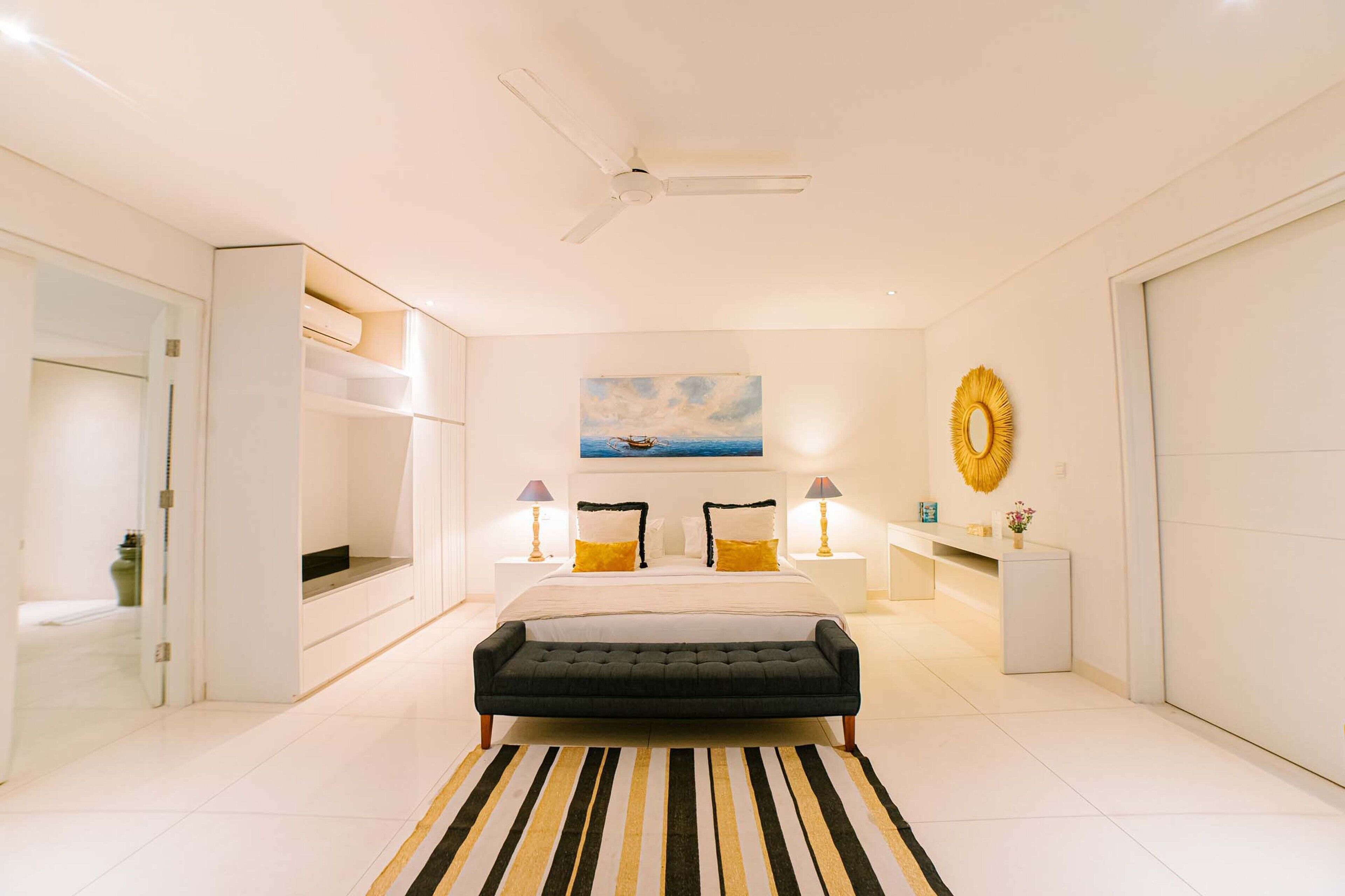A modern bedroom with a bed, bedside lamps, a ceiling fan, a painting, and a bench.