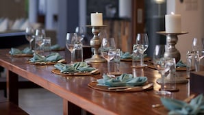 Mesa de jantar de madeira comprida posta com vários copos, pratos e guardanapos, com velas e uma moderna lumin ária pendente acima.