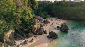 Uma praia com águas cristalinas, formações rochosas e algumas pessoas. Há árvores e um edifício ao fundo.