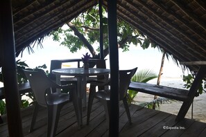 Un área de comedor al aire libre con techo de paja, mesa y sillas de madera, con vista a una playa.